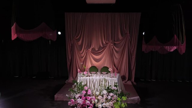 Luxurious wedding presidium table for the newlyweds standing in a dark, romantic banquet hall, beautifully decorated with pink curtains, fresh peonies, and elegant tableware for the celebration