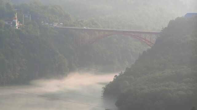 Cars driving on red bridge over Ena Gorge, Enakyo, Kiso River, Gifu Prefecture, Japan in the morning with mist underneath.
