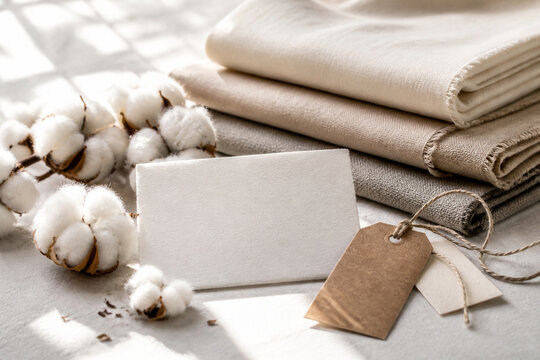 Soft neutral linen textiles stacked beside cotton bolls, a blank fabric swatch and kraft paper tags in warm sunlight.