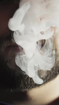 Extreme close-up of a bearded man's mouth and nose while he is vaping. He skillfully blows a thick cloud of vapor and smoke rings