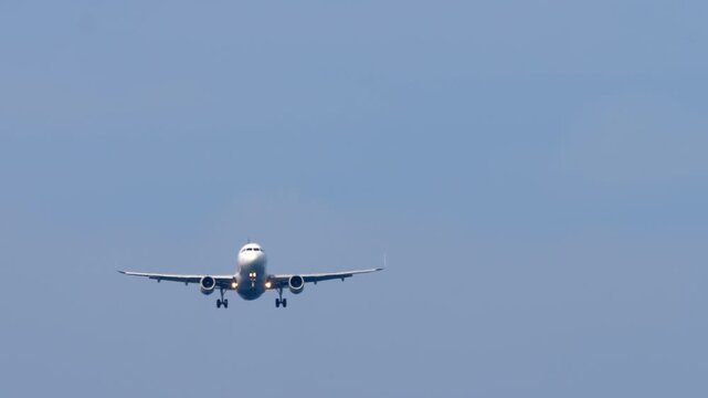 Passenger airplane flying head-on and descending for landing with gear down in clear blue sky during bright day. Telephoto shot with landing lights on.