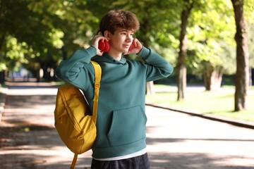 Naklejka premium Student with yellow backpack putting on headphones in park on sunny day