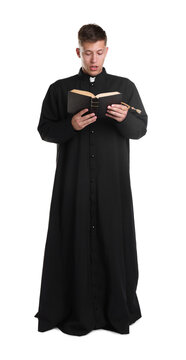 Young priest in cassock reading Holy Bible on white background