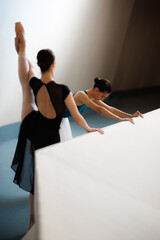 Two ballet dancers stretching at a barre during rehearsal, one in black leotard lifting leg while partner leans forward in teal showing focus and graceful technique © Nikola Spasenoski