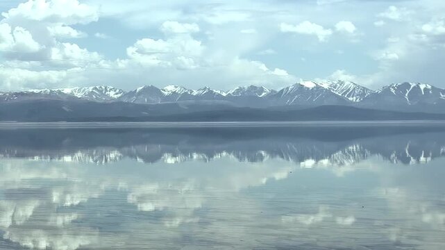 Experience the breathtaking tranquility of Khuvsgul Lake in Mongolia. Pristine waters perfectly mirror a vast sky adorned with fluffy clouds and the majestic silhouette of snow-capped mountains.
