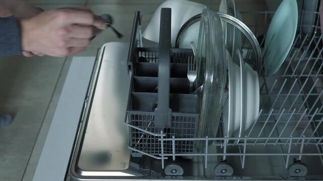 Close front view inside open dishwasher showing hand placing utensil into basket while plates, glasses, and dishware settle upright in lower rack slots within stainless steel kitchen appliance
