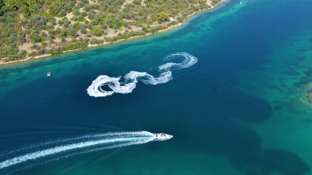 Aerial view of a jet ski speeding across deep blue Mediterranean water, creating a long white wake while riding through a scenic turquoise bay surrounded by green coastal hills.