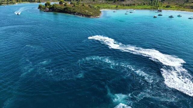 Aerial view of a jet ski speeding across deep blue Mediterranean water, creating a long white wake while riding through a scenic turquoise bay surrounded by green coastal hills.