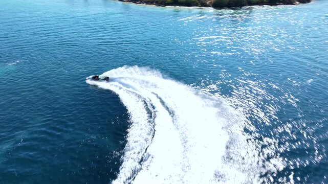 Aerial view of a jet ski speeding across deep blue Mediterranean water, creating a long white wake while riding through a scenic turquoise bay surrounded by green coastal hills.