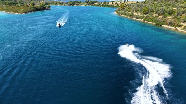 Aerial view of a jet ski speeding across deep blue Mediterranean water, creating a long white wake while riding through a scenic turquoise bay surrounded by green coastal hills.