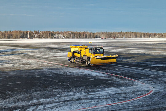 Airport snow removal truck clearing icy airport surface, snow-covered airfield and forest in background during bright winter day: Riga, Latvia - January 13, 2025