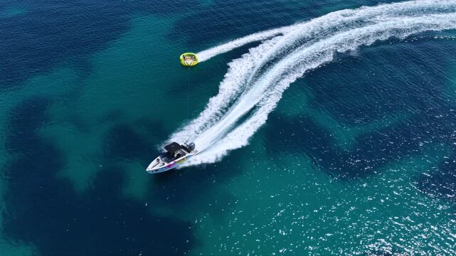 Aerial view of a speedboat towing an inflatable tube across deep blue sea near a sandy beach and coastal resort in Sithonia, Halkidiki, Greece, creating a long white wake on a sunny summer day.