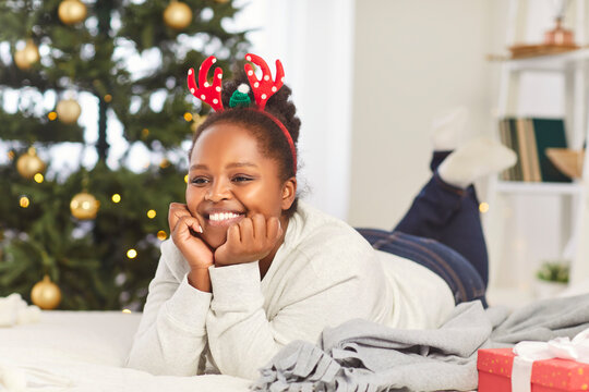 Portrait of a smiling African American woman lying on the floor beside a Christmas tree at home. She celebrates New Year or Christmas morning with happiness and warmth in a cozy holiday atmosphere.