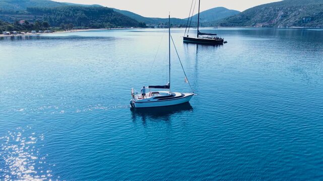 Aerial view of a large sailing yacht anchored with its tender boat in a calm sheltered bay in Sithonia, Halkidiki, Greece, surrounded by clear blue water, sandy beach and forested hills.