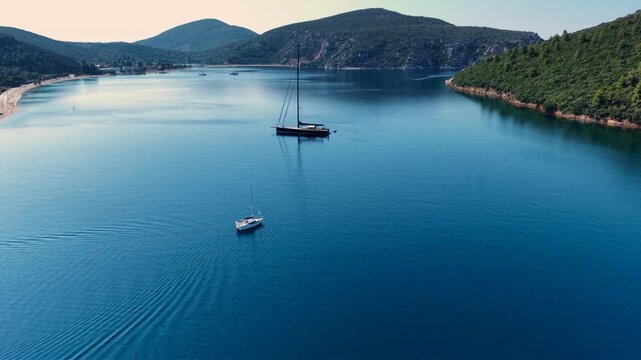 Aerial view of a large sailing yacht anchored with its tender boat in a calm sheltered bay in Sithonia, Halkidiki, Greece, surrounded by clear blue water, sandy beach and forested hills.