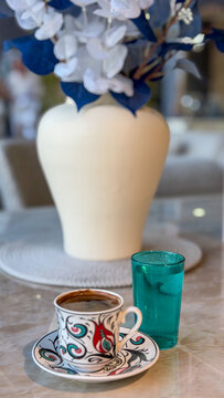 Traditional Turkish coffee served in a decorative ceramic cup with a glass of water, elegant floral arrangement and modern interior in the background.