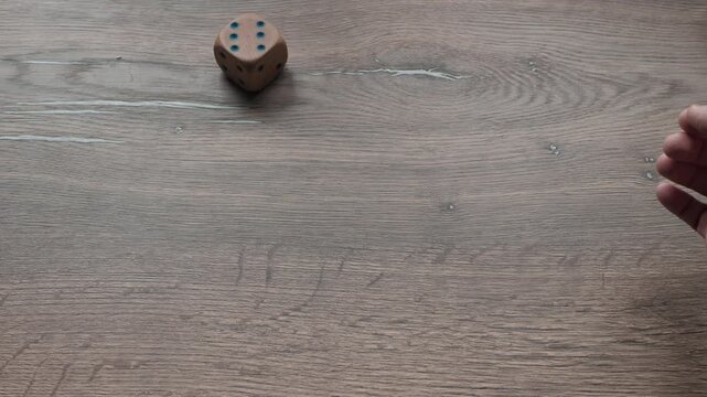 Close view showing right male hand releasing wooden six sided die on rustic textured tabletop, die rolling across surface then settling while hand pulls back