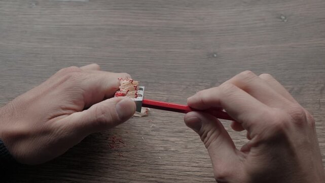 Top-down view of two hands sharpening red pencil with handheld metal sharpener on warm wooden table, with curled shavings, steady grip, and clear close detail of simple everyday desk work