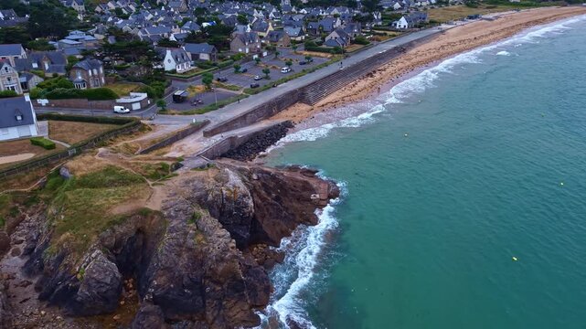 Approaching drone movement over a rocky coastal headland near Saint-Lunaire residential buildings overlooking Atlantic waves at Longchamp Beach with dam and footpath, Cote d'Emeraude, Brittany, France