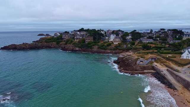 Approaching drone fly near Longchamp Beach toward coastal Saint-Lunaire residential neighborhood surrounding bay environment and rocky shoreline, Cote d'Emeraude, Brittany, France