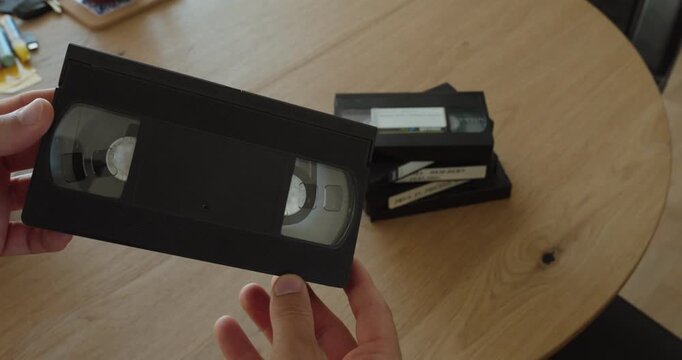 Close-up of hands holding and examining an old VHS cassette with more videotapes in the background. Ideal for vintage technology, analog video, nostalgia, home movies, archives, and media digitization