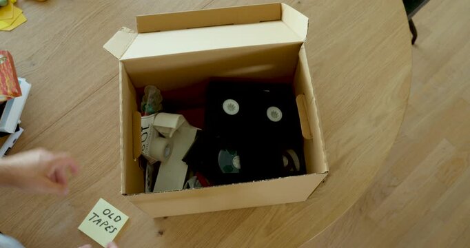 Person putting old VHS tapes into a cardboard box for storage. Ideal for nostalgia, old memories, archiving, decluttering and retro technology.