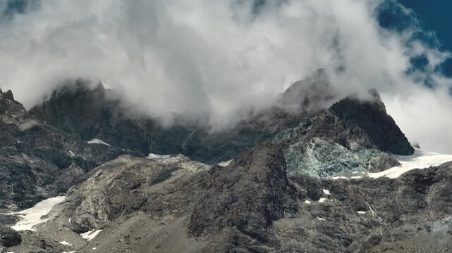 Close-up of dramatic alpine mountain peaks wrapped in rolling clouds, revealing lingering glacier patches between rugged summits beneath changing mountain weather.