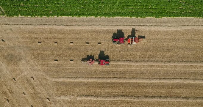 Top-down drone shot of two red tractors driving toward each other while baling straw on a harvested wheat field with perfectly parallel windrows in the Dutch countryside.