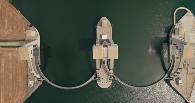 Top-down drone ascends to reveal the full complex of a hydroelectric power plant and its distinctive arch dam in a Dutch canal, highlighting renewable energy infrastructure.
