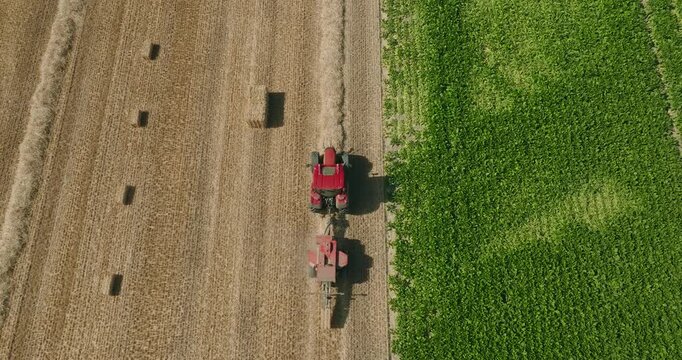 Top-down drone follows a tractor towing a straw baler over harvested wheat windrows as rectangular straw bales are ejected across a sunny agricultural landscape.