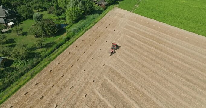 Wide aerial drone shot approaching a tractor towing a straw baler as it ejects rectangular straw bales across a harvested wheat field surrounded by lush green farmland on a sunny day.