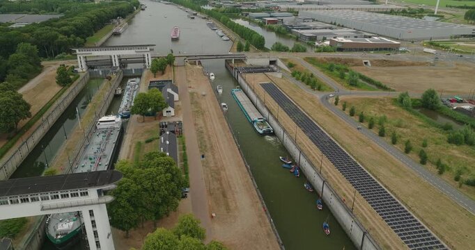 Drone flies over a canal sluice with two tanker barges waiting in the lock, while leisure boats pass in the channel next the lock, with solar panels on the shore, windmills and a business park beyond.