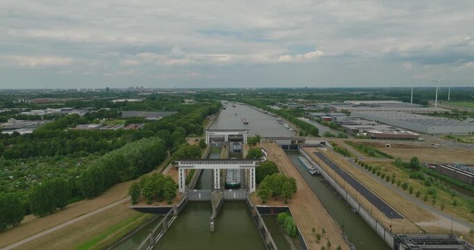 Drone approaches a closed sluice gate with an oil tanker barge waiting in the lock chamber, surrounded by Dutch polder countryside, a major river, commercial shipping and a provincial road.