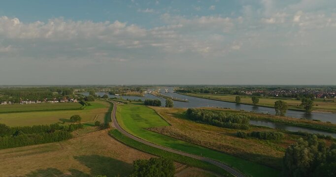Beautiful aerial view of a sunny Dutch summer polder landscape with a wide river, tree-lined riverbank, grassy dyke and flat countryside beneath a clear blue sky.