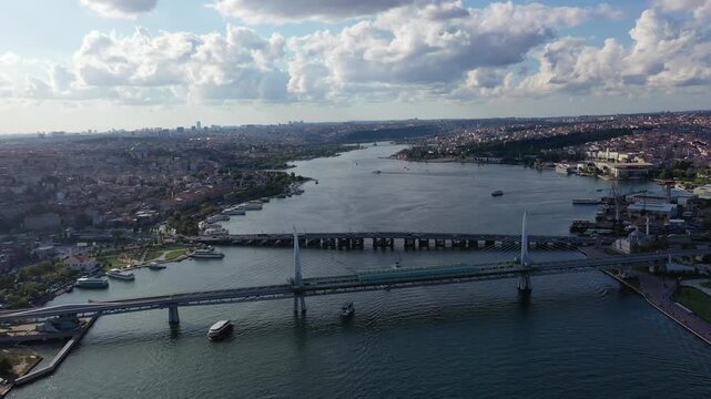 High aerial drone shot of the Golden Horn Metro Bridge and Atat&uuml;rk Bridge spanning the water with boats passing below and the Istanbul cityscape stretching toward a cloudy sky in Turkey.