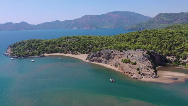High aerial drone view of a secluded sandy cove with turquoise clear water, rocky cliffs, anchored boats, and pine tree-covered hills along the Mediterranean coast in Mugla, Turkey.