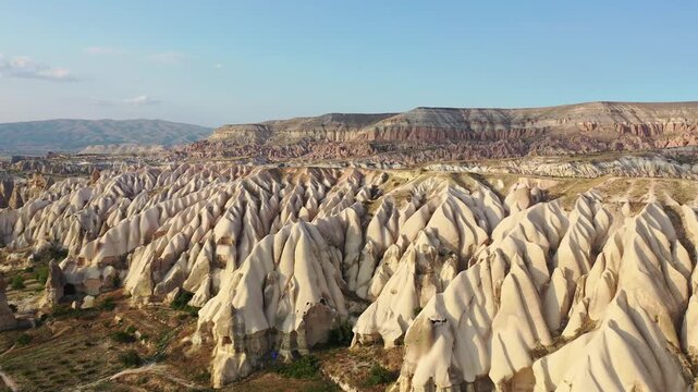 Panoramic aerial drone view of white rock formations, conical fairy chimneys, and flat-topped tuff mountains in Rose Valley, Cappadocia, Nevsehir, Turkey under a clear blue sky.