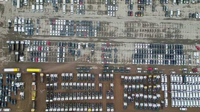 Top-down aerial shot of a large industrial vehicle distribution center showing perfectly aligned rows of parked cars and yellow tractors.