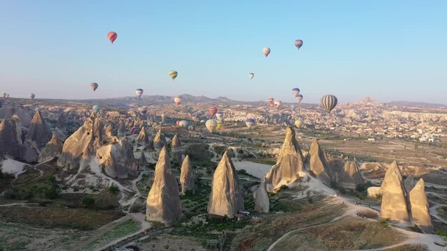 Wide aerial drone view of multiple hot air balloons floating across the sky over the dramatic valley landscape of Cappadocia, Nevsehir, Turkey at sunrise.