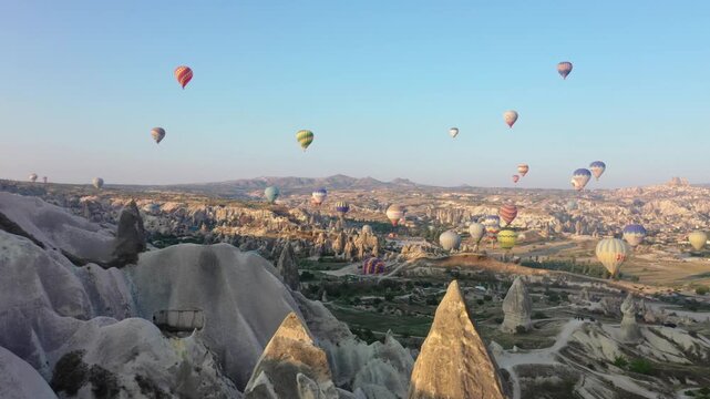 Wide aerial drone view of multiple hot air balloons floating across the sky over the dramatic valley landscape of Cappadocia, Nevsehir, Turkey at sunrise.