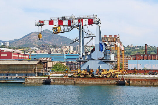 Genoa, Italy - 29 August 2018: View of the industrial area of Genoa Harbor featuring a large red and white crane, industrial buildings, and hills in the background under a clear sky.