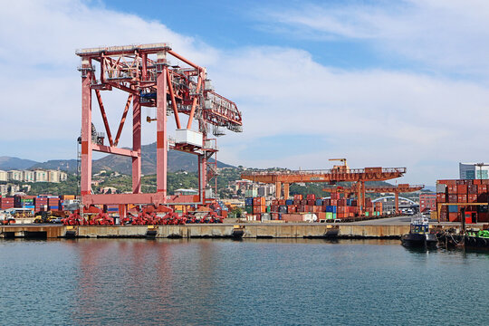 Genoa, Italy - 29 August 2018: View of the Genova Harbor industrial area with large red container cranes, stacks of colorful shipping containers, and calm harbor waters.