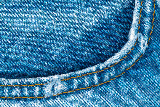 Macro detail of classic blue denim jeans fabric. Worn texture with frayed front pocket edge and contrast orange stitching creates simple, casual background for fashion or textile concepts