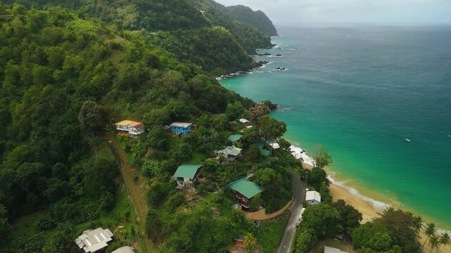 Aerial parallax reveals Castara Bay in Tobago with colorful eco lodges nestled in rainforest winding cliffside road and secluded turquoise beach cove