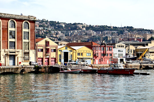 Genoa, Italy - 29 August 2018: View of Genova Harbor industrial area with colorful warehouses, docked patrol boats, and the hillside cityscape in the background.