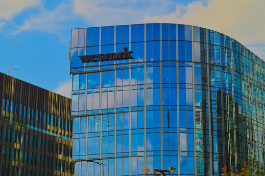 Modern glass WeWork coworking office building exterior with blue reflective windows against cloudy blue sky in urban city district