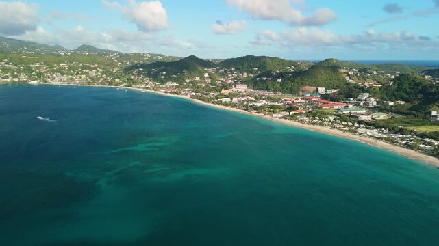 High altitude aerial pans across turquoise Grand Anse Bay in Grenada revealing sandy beach town and green mountain backdrop