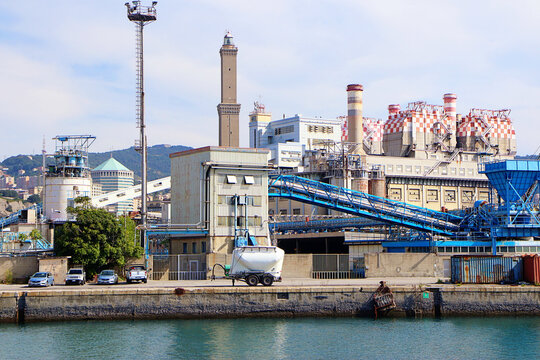 Genoa, Liguria, Italy - 29 August 2018: View of the Lanterna lighthouse overlooking the industrial harbor area with large storage facilities, blue sky, and calm water.
