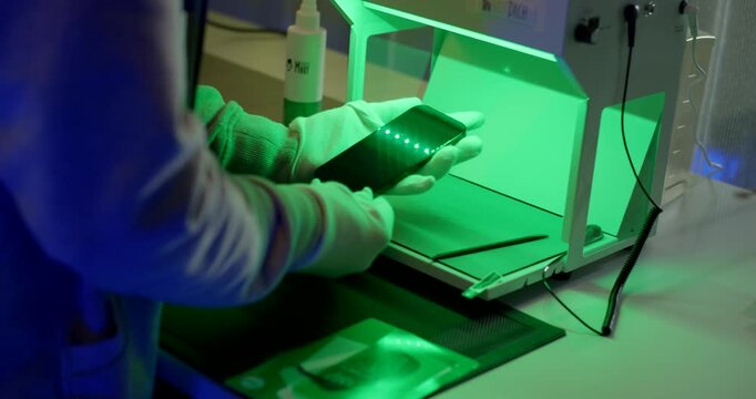 Gloved hand inspecting smartphone screen under green light booth