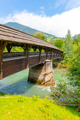 Fototapeta premium A long, covered wooden bridge spans a clear mountain stream surrounded by lush green foliage in a serene valley. Austria